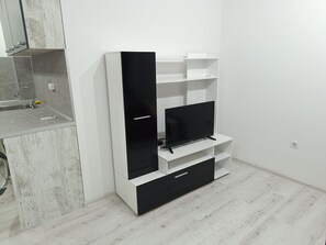A modern living room with a flat-screen TV, built-in shelves, and a kitchen area with a sink and countertop. - One-Bedroom Apartment-14 Filip Makedonski 49B (Plovdiv)