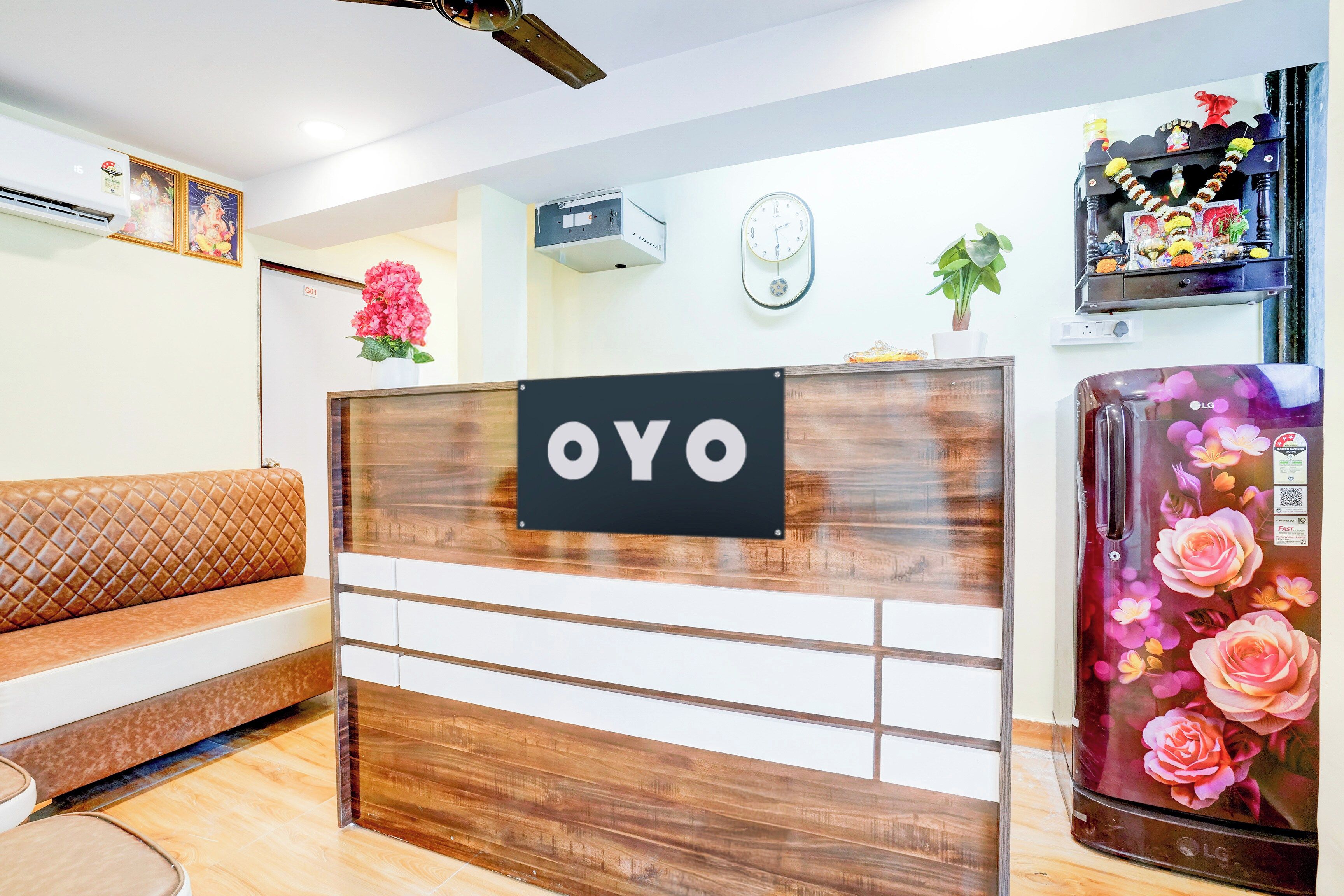 Un mostrador de recepción con un letrero que dice 'OYO', un reloj, una planta en maceta y un refrigerador con flores.