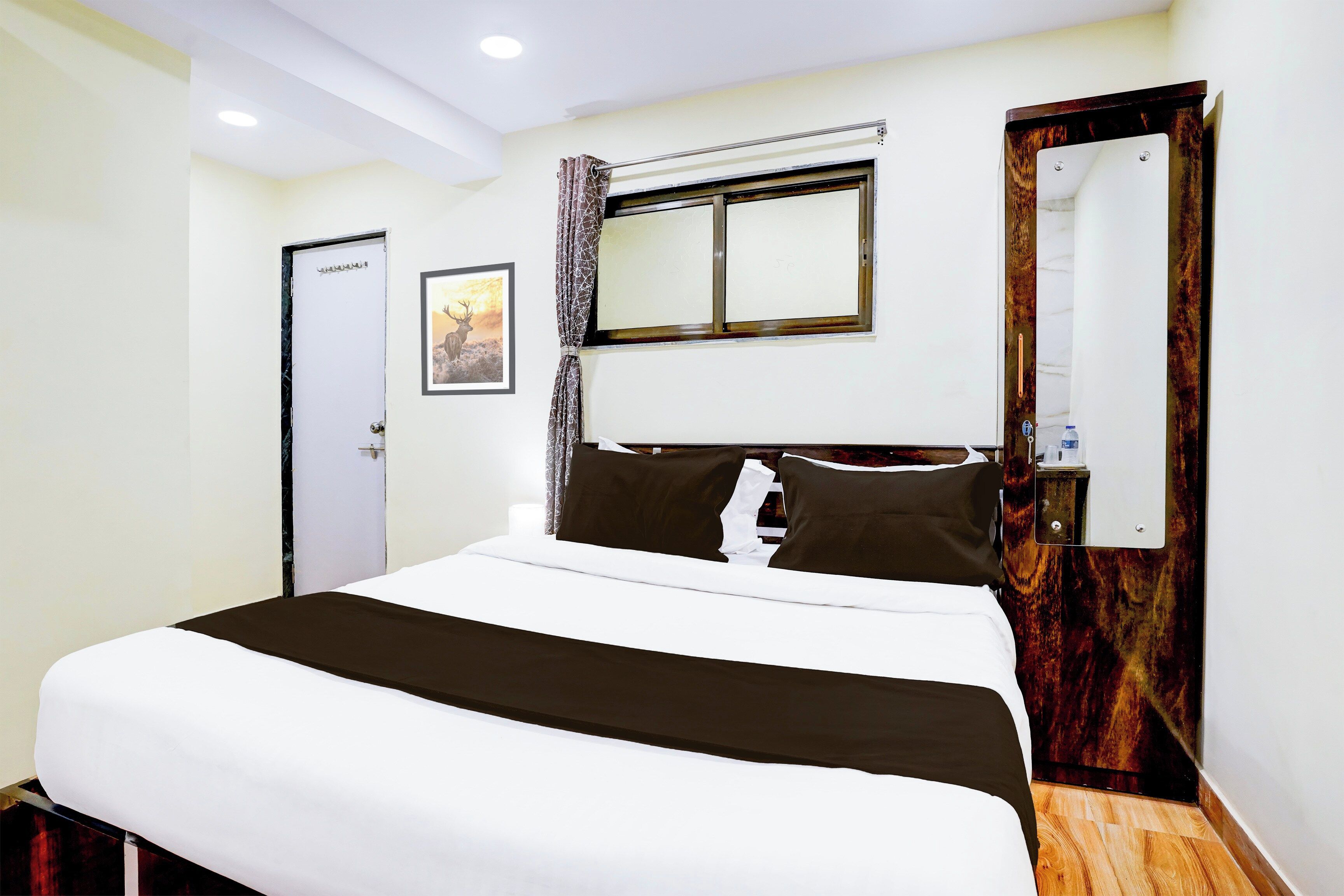 Habitación de hotel con cama, puerta, ventana, espejo y un cuadro colgado en la pared.