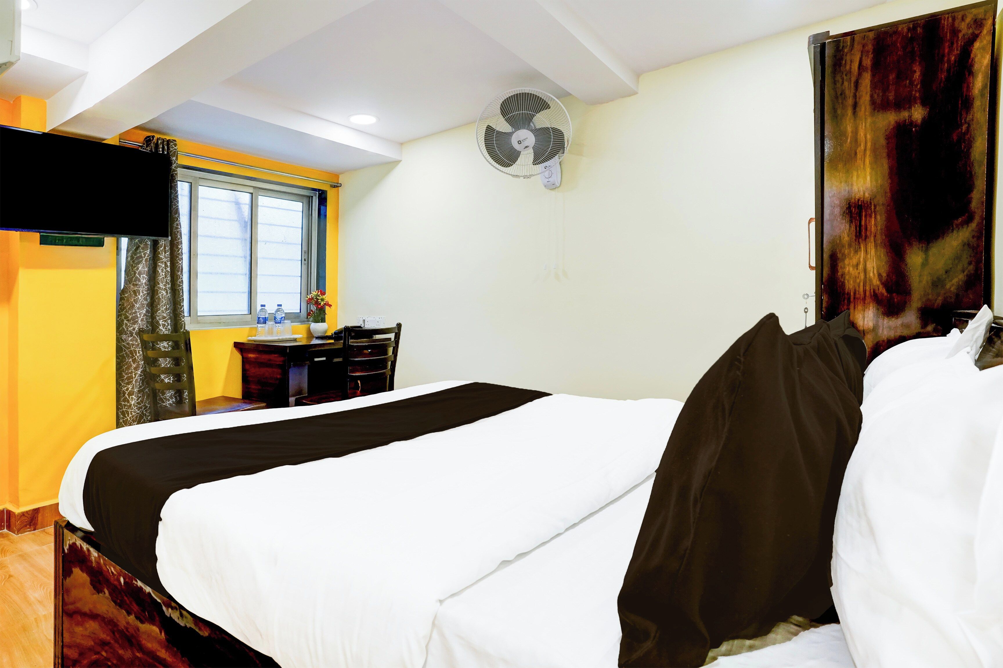 Una habitación de hotel con cama, escritorio, silla, ventana y ventilador de pared.