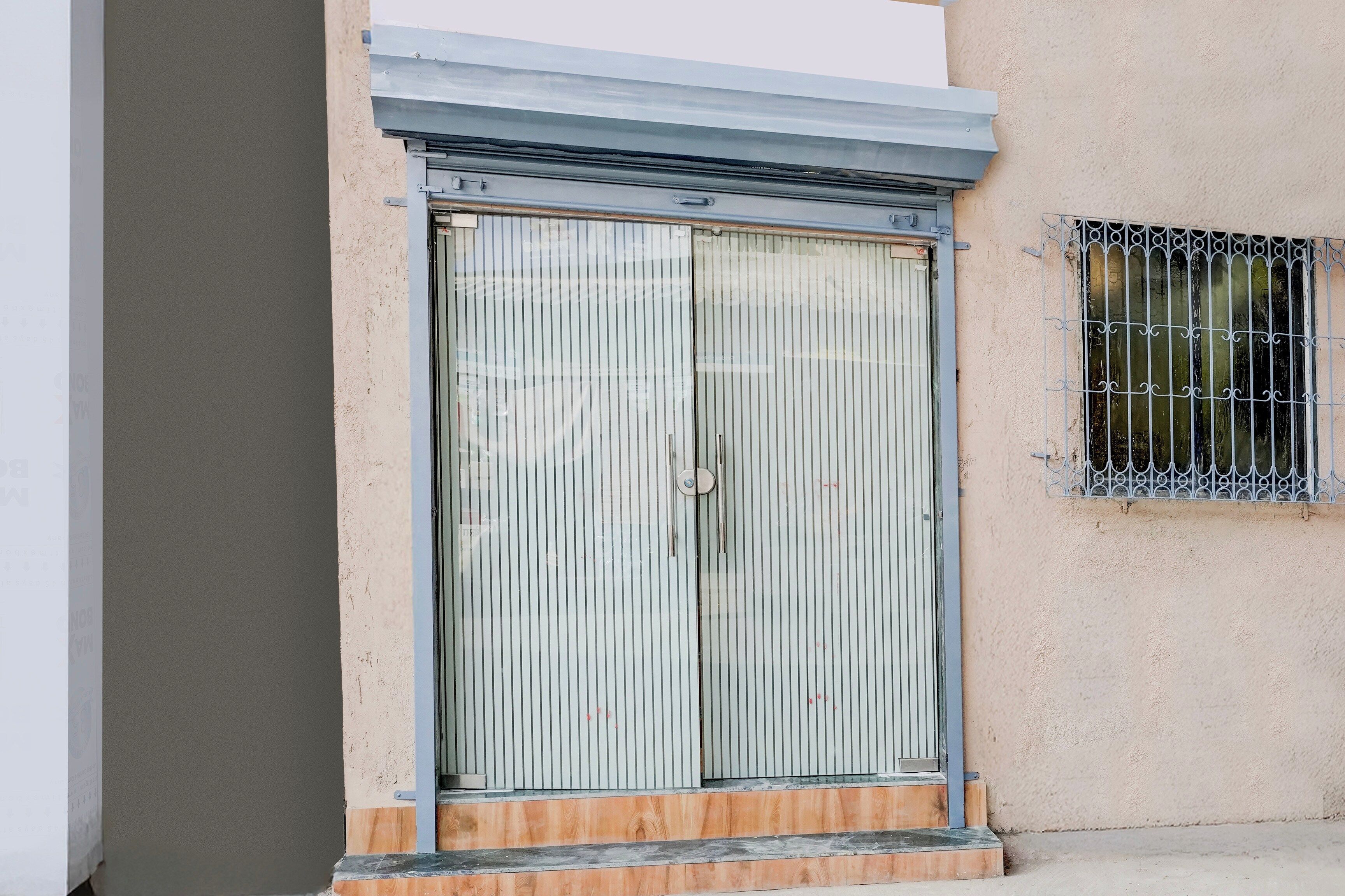 Puertas dobles con persianas verticales, un ventanal pequeño con barandillas decorativas y un escalón de hormigón que lleva a la puerta.