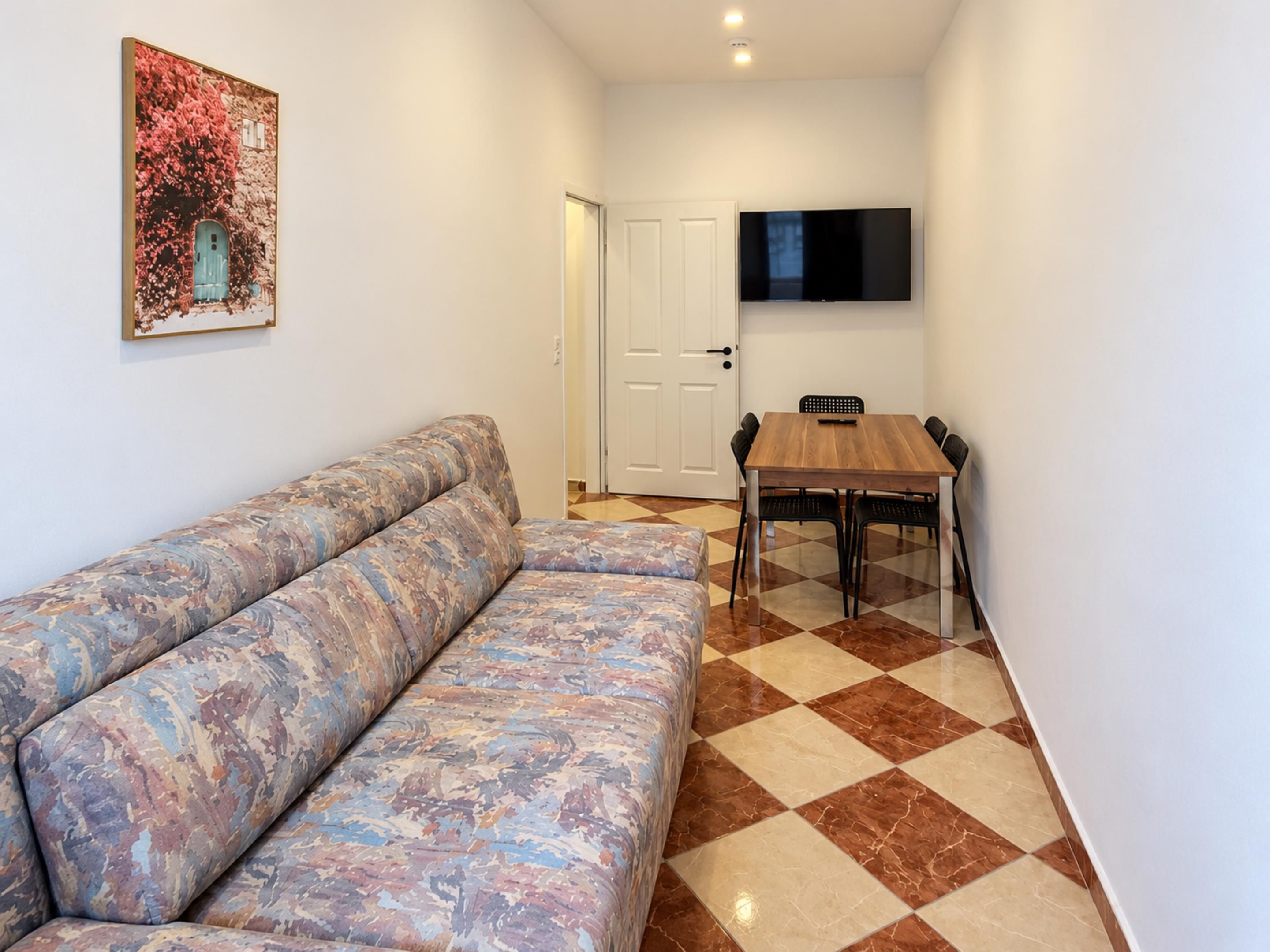 Een zithoek met een bank, een eettafel met stoelen, een flatscreen televisie aan de muur en een deur naar een andere kamer.