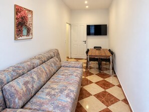 Een zithoek met een bank, een eettafel met stoelen, een flatscreen televisie aan de muur en een deur naar een andere kamer.