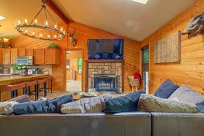 A cozy cabin interior with a stone fireplace, wooden walls, a bar area, and a seating area with cushions. - Walk to Midtown! Family Getaway w/ Wraparound Deck (Ruidoso)