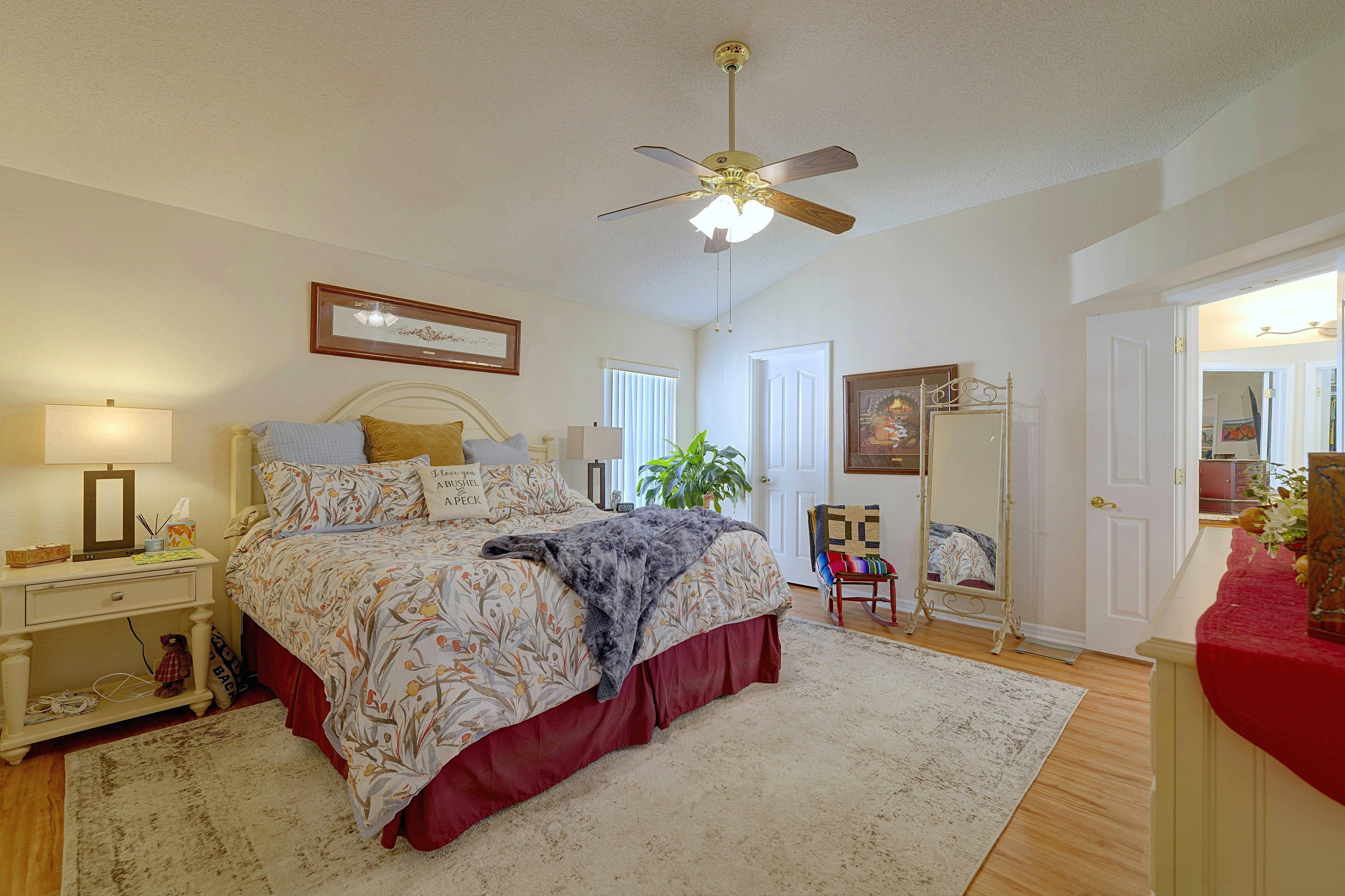 A bedroom with a bed, bedside table, nightstand, lamp, ceiling fan, and a door leading to another room.