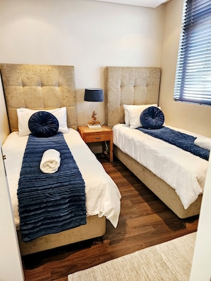Ein Hotelzimmer mit zwei Betten, einem Nachttisch mit Lampe und einem Fenster mit Jalousien.