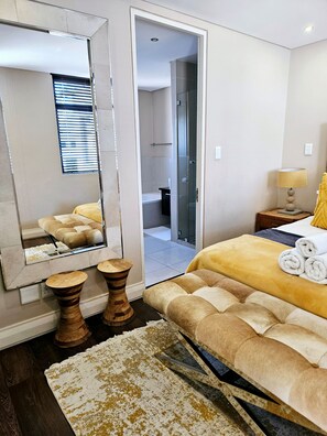 A modern bedroom with a large mirror, a bed with rolled towels, a bedside table with a lamp, and a view into the bathroom. - Lovely 2-bedroom apartment in Big Bay for a relaxing stay (Cape Town)