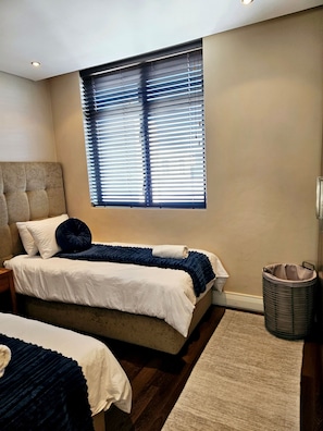 Une chambre à coucher avec un lit, une fenêtre avec des stores, une table de chevet et une corbeille à déchets.