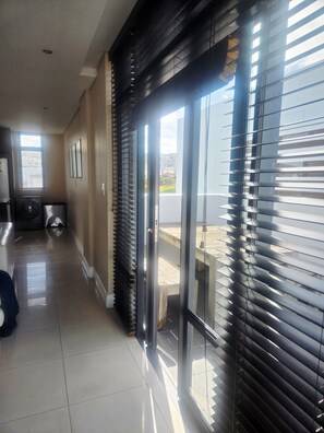A corridor with a washing machine, a door with blinds, and a view of buildings outside. - Lovely 2-bedroom apartment in Big Bay for a relaxing stay (Cape Town)