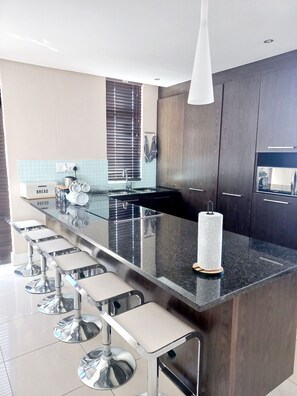 A modern kitchen with a central island, bar stools, and a built-in oven. - Lovely 2-bedroom apartment in Big Bay for a relaxing stay (Cape Town)