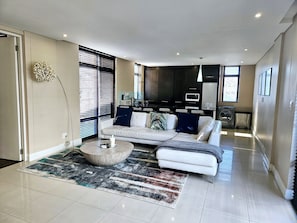 A modern living room with a sofa, coffee table, and a kitchen area in the background. - Lovely 2-bedroom apartment in Big Bay for a relaxing stay (Cape Town)