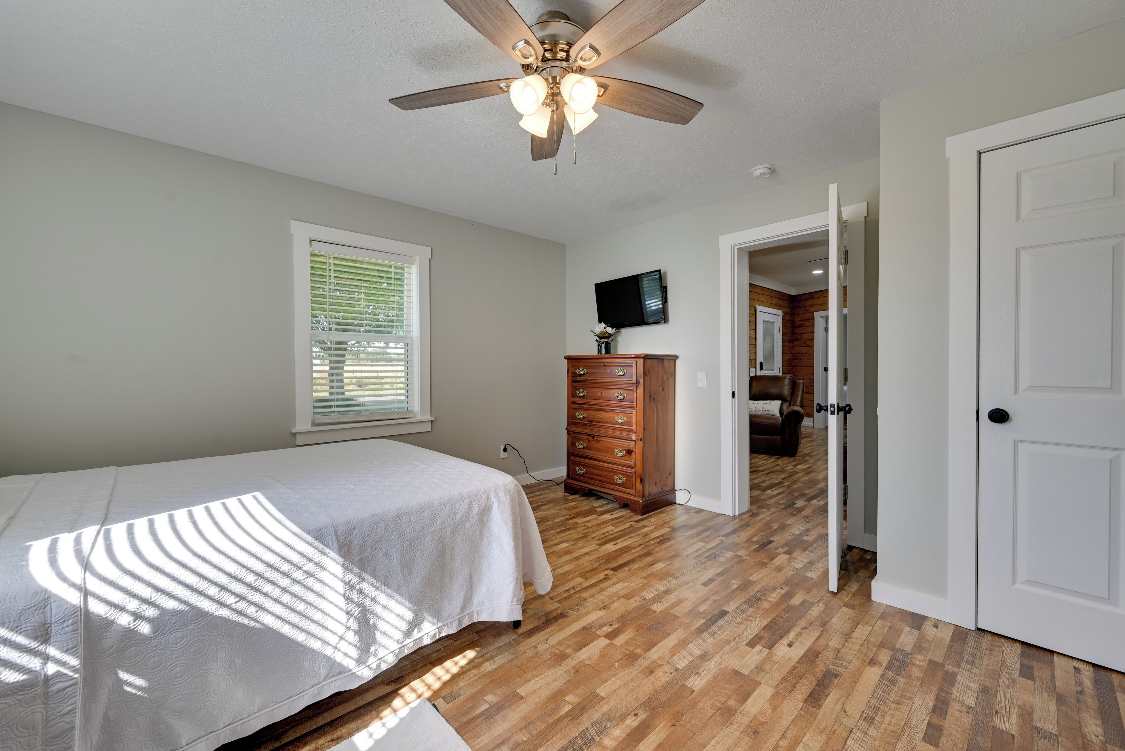 A bedroom with a bed, wooden dresser, ceiling fan, and a television on the wall.