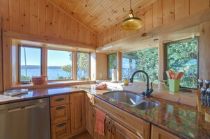 Una cocina con armarios de madera, encimera de granito, un lavamanos y ventanas amplias con vistas al exterior.