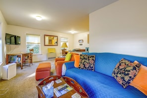 A living room with a blue sofa, a wooden coffee table, a TV mounted on the wall, and a window with blinds. - Saint Albans Home: Walk to Main Street! (St. Albans)