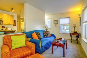 A living room with a blue sofa, orange armchair, and a wooden coffee table. There is a kitchen area in the background with wooden cabinets and a window.