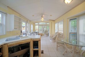 A kitchen with a central island, white cabinets, and a dining area with wicker chairs and a glass table. - Water Views + Patio Brews! Middle Bass Island Home (Put-in-Bay)