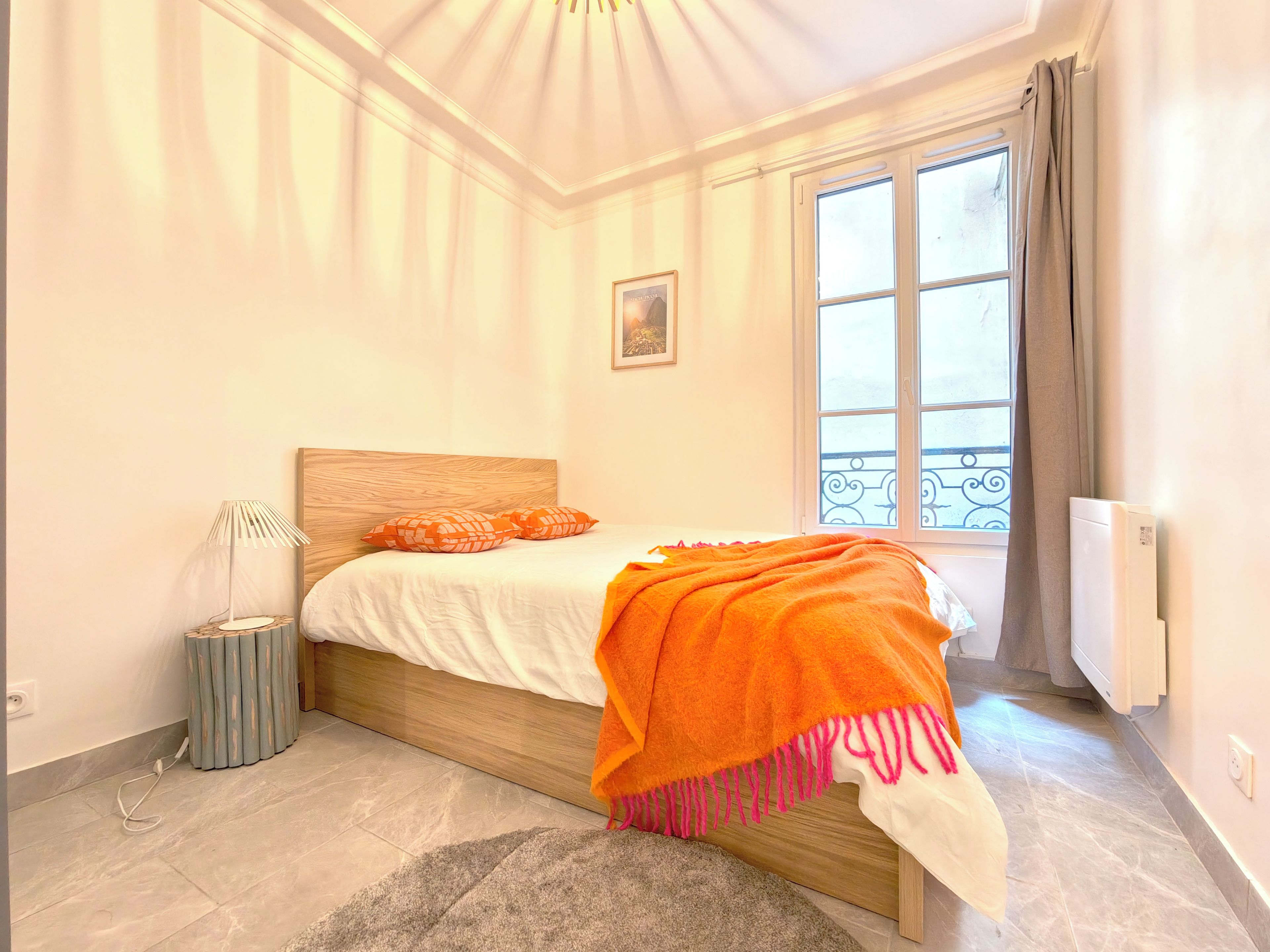Un dormitorio con cama, mesita de noche, lámpara, radiador y ventana con cortinas.