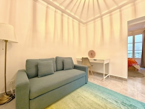 Ein Zimmer mit grauem Sofa, Schreibtisch mit Stuhl, Lampe und einem Fenster mit Vorhängen.