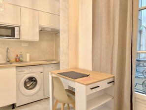 Eine kleine Küche mit Waschmaschine, Mikrowelle und Spüle. Ein kleiner Tisch mit Stuhl und ein Fenster mit Vorhängen.