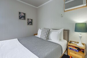 Una cama bien tendida con una colcha gris estampada, un almohadón blanco y otro a cuadros. En la pared, dos cuadros con motivos florales. Se ve una mesita de noche con una lámpara y algunos libros.