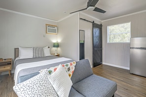 A modern bedroom with a bed, a sofa, a desk, a lamp, a ceiling fan, a window, and a refrigerator. - Boathouse Cabin at Horse Creek Retreat (Fort Ogden)