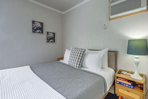 Ein ordentlich bezogenes Bett mit einer grauen, gemusterten Tagesdecke, einem weißen Kissen und einem karierten Kissen. An der Wand hängen zwei gerahmte Blumenbilder. Ein Nachttisch mit einer Lampe und einigen Büchern ist zu sehen.