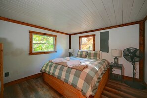 Ein Schlafzimmer mit einem hölzernen Bett, zwei Fenstern, einem Standventilator und Holzboden.