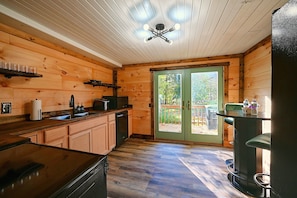 A wooden cabin interior with a kitchen area, a dining table, and a sliding glass door leading to an outdoor deck. - Hocking Hills Area Retreat: Relax & Explore! (Rockbridge)