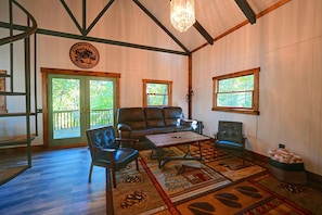 Ein geräumiges Zimmer mit einem großen Ledersofa, einem Couchtisch und einem Teppich mit Waldmotiv. Zwei Fenster sorgen für natürliches Licht und eine Tür führt nach draußen.