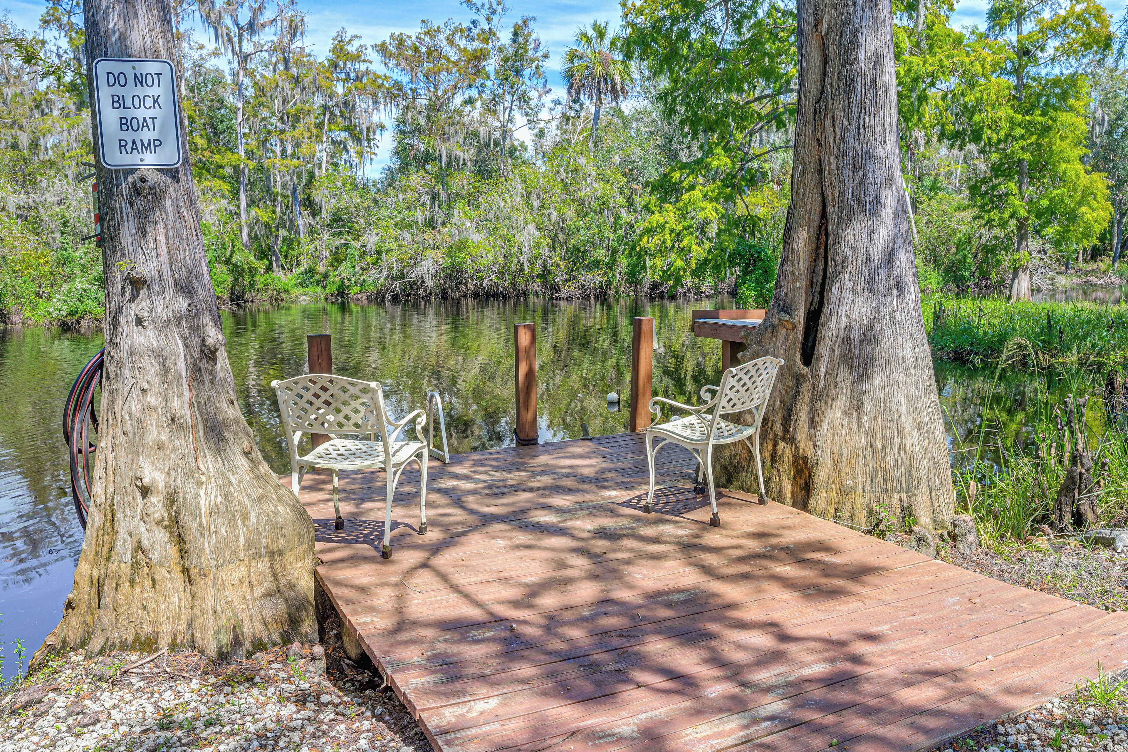 A wooden dock with two white metal chairs, surrounded by trees and a body of water.