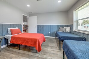 A room with two beds, one with a red bedspread and the other with a blue bedspread, a window with blinds, and a door leading to another room. - Riverside Retreat -horse Creel Retreat Arcadia (Fort Ogden)