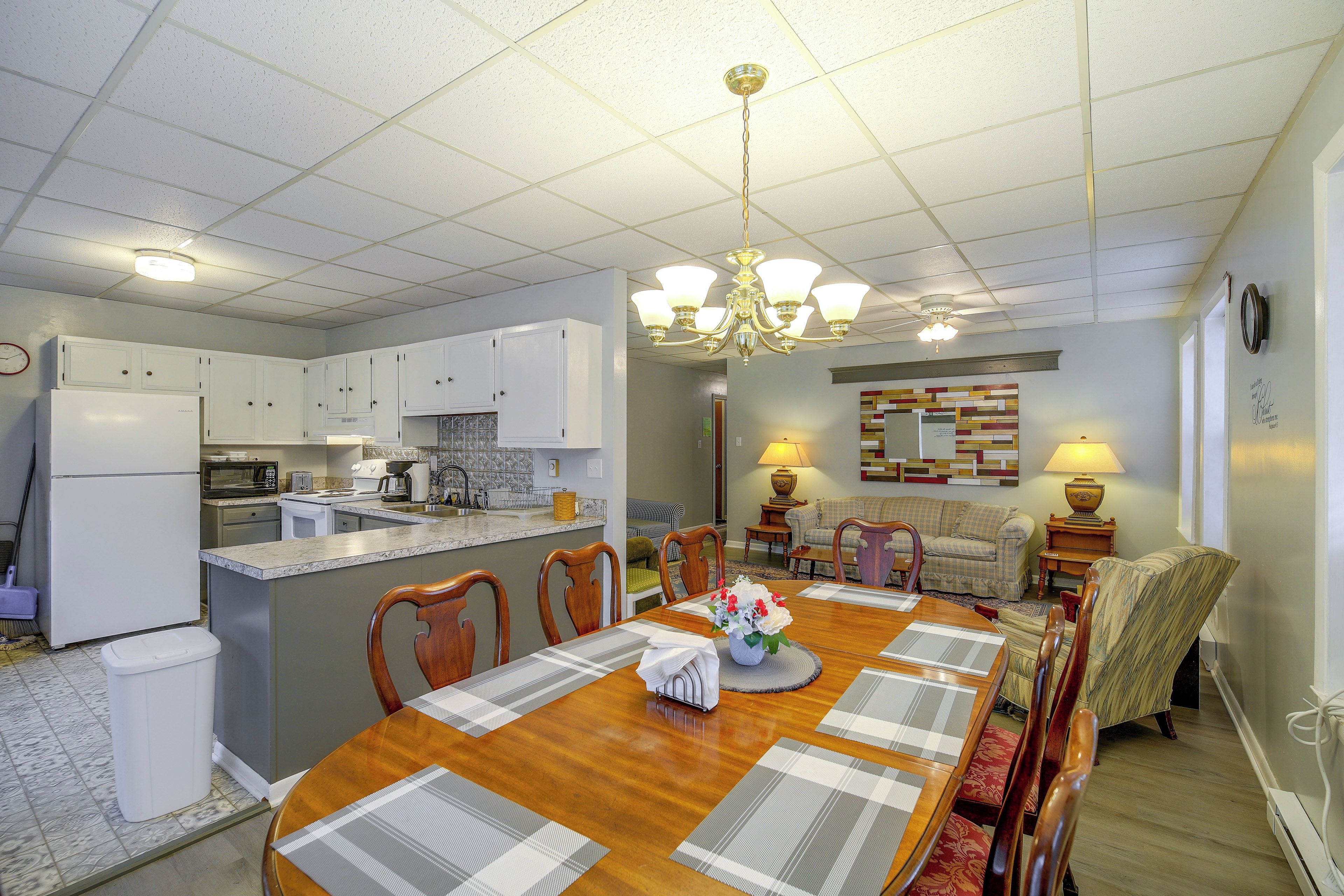 A dining area with a wooden table, chairs, a chandelier, and a kitchen in the background.