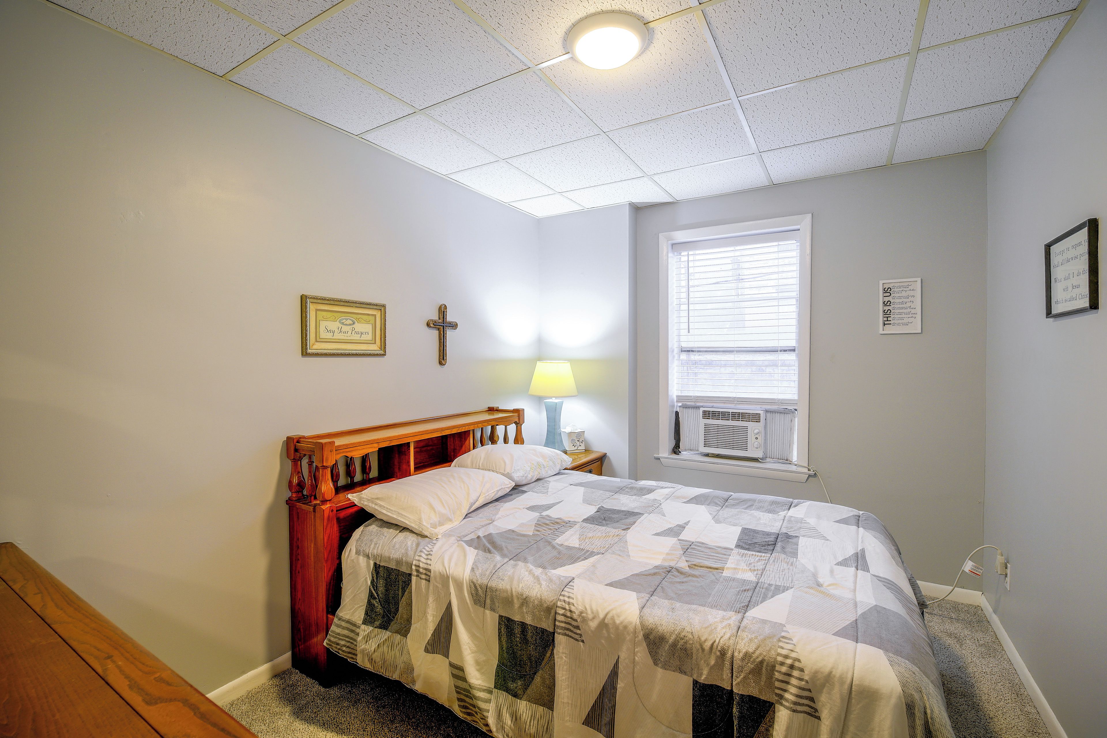 A bedroom with a wooden bed, a nightstand, a cross on the wall, and a window with blinds.