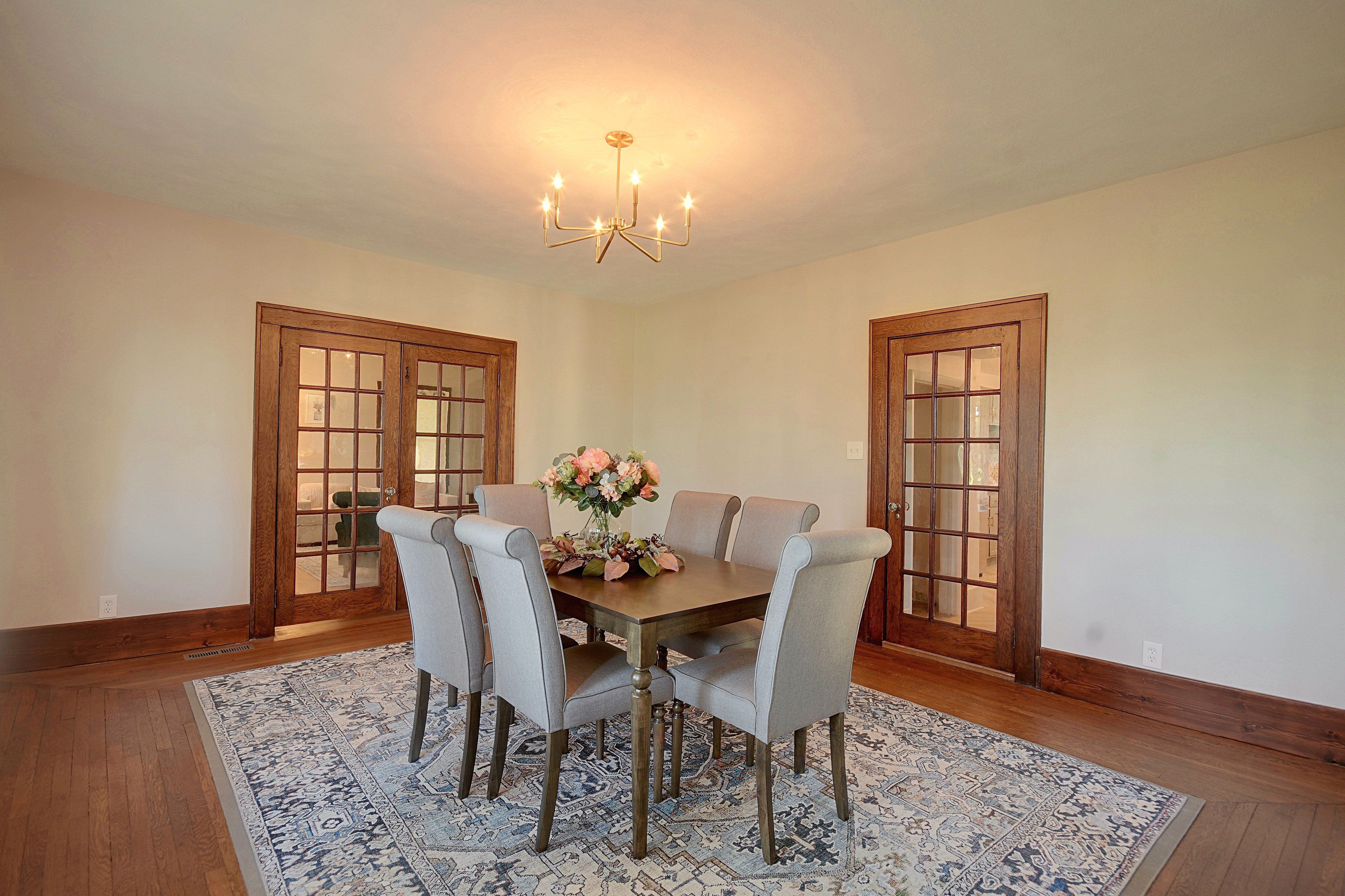 A dining room with a wooden table, six chairs, a chandelier, and a floral centerpiece.