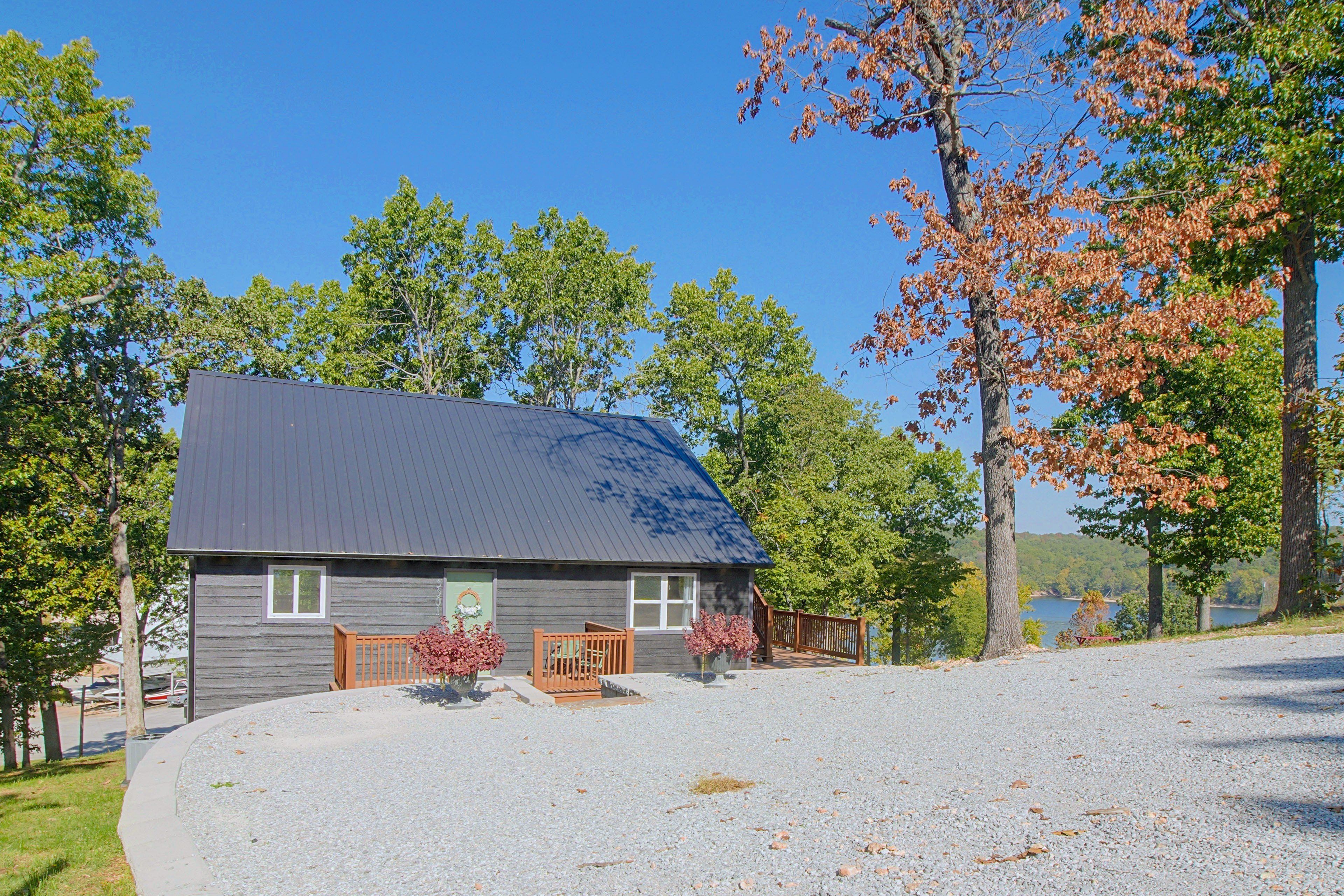 A two-story cabin with a gravel driveway, surrounded by trees and a lake view.