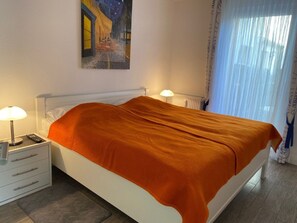 A hotel room with a bed, a nightstand with a lamp, a painting on the wall, and a window with curtains. - South Beach Room (Wyk auf Foehr)