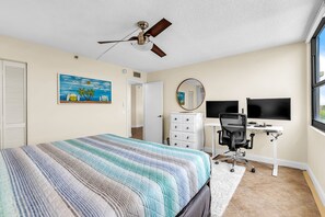 A bedroom with a bed, a desk with a computer, a chair, a dresser, a ceiling fan, a painting, and a window. - Water Views/Pool - Bahia Shores O-503 - Sunrise on the Bay (St. Petersburg)