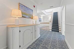 Un couloir avec une console blanche, deux lampes et un tableau accroché au mur. Un escalier recouvert d’une moquette bleue mène à un palier.