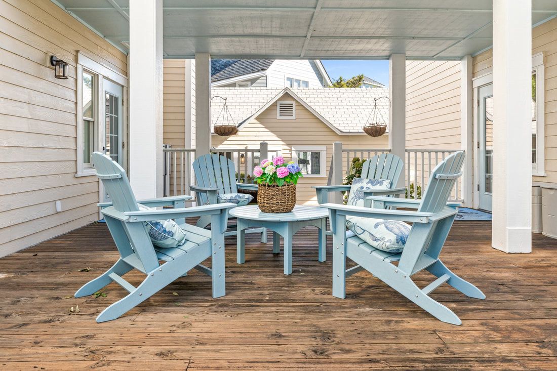A covered porch with light blue Adirondack chairs, a central table with a basket and flowers, and a house in the background.