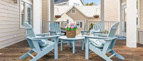 A covered porch with light blue Adirondack chairs, a central table with a basket and flowers, and a house in the background.