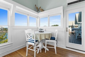 Un comedor con una mesa redonda y sillas blancas, ventanas amplias con vistas al agua y edificios, y una puerta que da a una terraza.