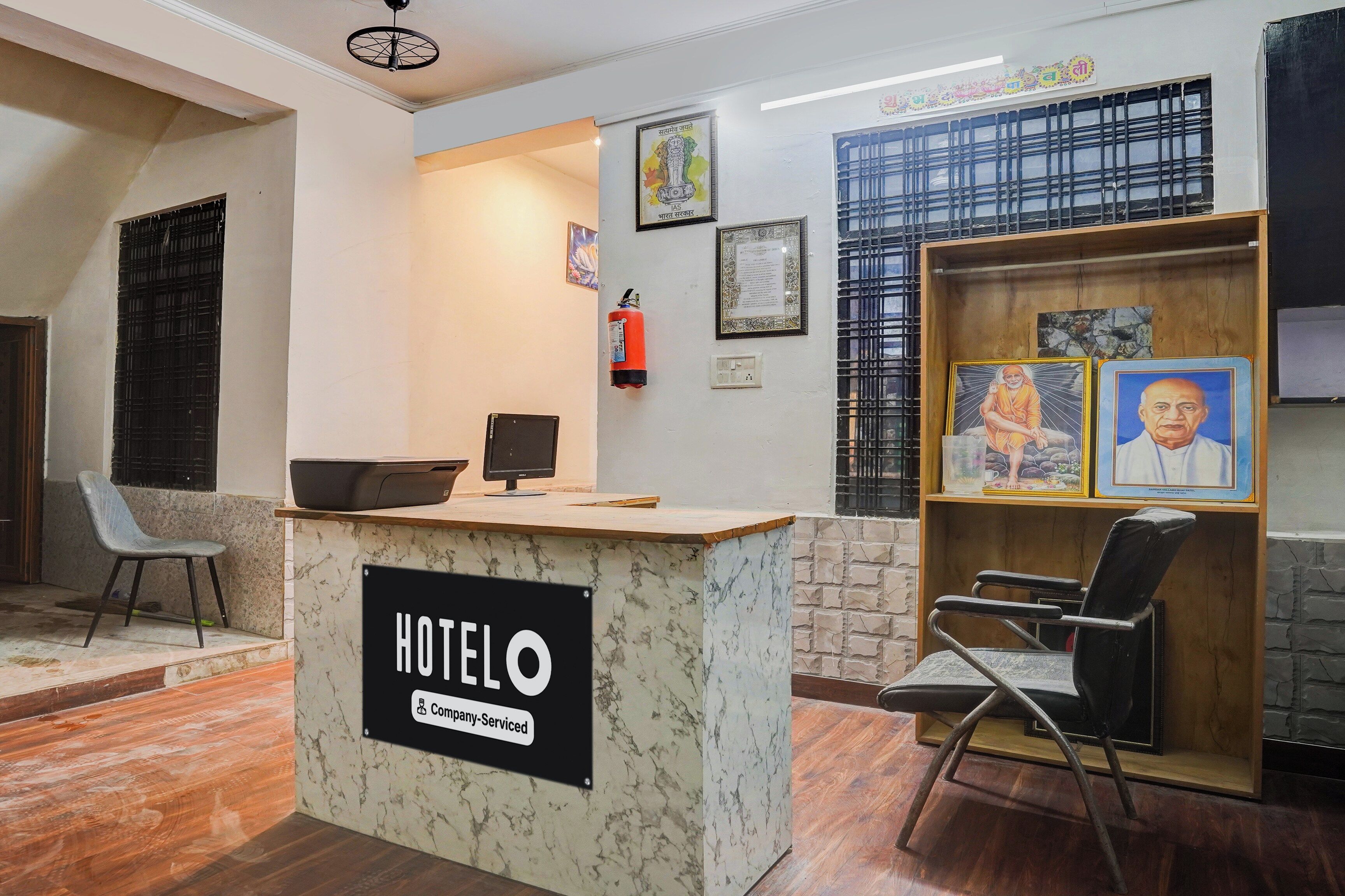 A hotel reception desk with a sign indicating it is company-serviced. There are framed pictures on the wall, a fire extinguisher, and a window with metal bars.