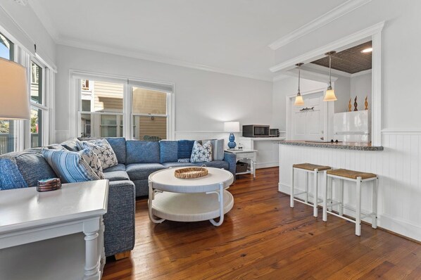 A modern living room with a sectional sofa, a round coffee table, and a kitchen area with a white countertop and bar stools. - This enchanting handicap accessible 1st floor suite is a perfect gateway to enjo (Bald Head Island)