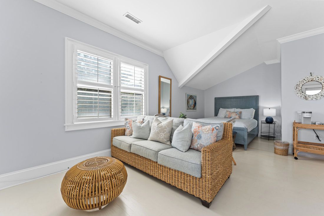 A bedroom with a sloped ceiling, a bed, a sofa, a round wicker ottoman, and a bedside table with a lamp.