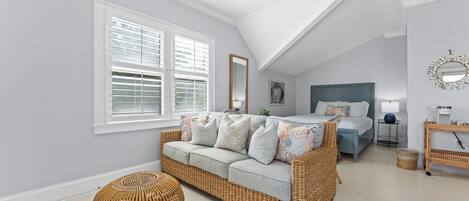A bedroom with a sloped ceiling, a bed, a sofa, a round wicker ottoman, and a bedside table with a lamp.