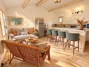A cozy living room with a sofa, a wicker chair, a coffee table, and a kitchen area with a bar and stools. - Bergerie Murtinella (Olmeto)