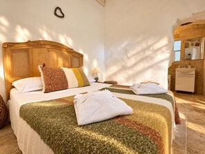 Quarto de hotel com cama de madeira, mesa de cabeceira, luminária e banheiro com pia e espelho.