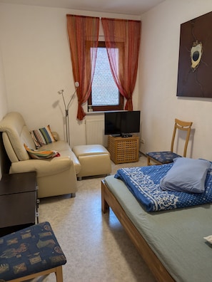 Ein Schlafzimmer mit einem Bett, einem Sessel, einer Couch, einem Fernseher und einem Fenster mit Vorhängen.