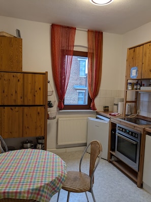 Eine Küche mit Fenster, Holzschränken, einem Tisch mit kariertem Tischläufer, einem Korbstuhl und einem eingebauten Backofen.