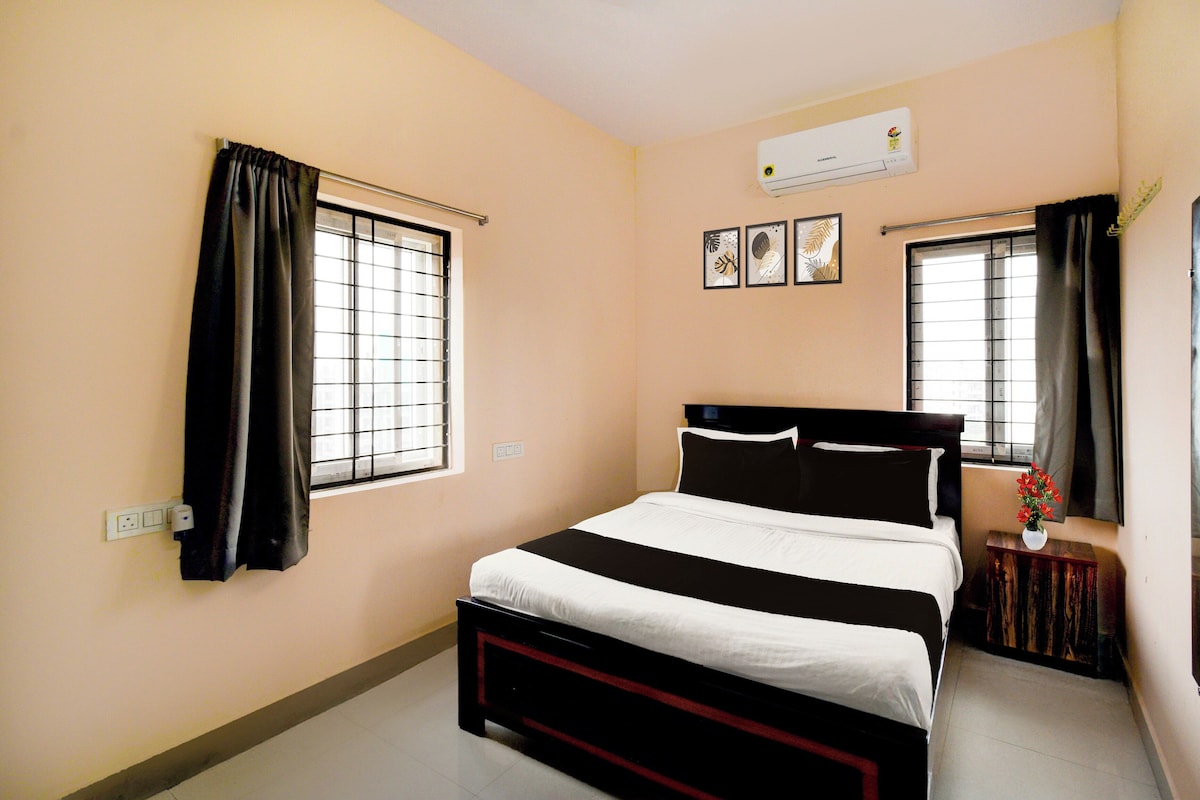 A bedroom with a bed, two windows with curtains, an air conditioner, and a small bedside table with a vase.